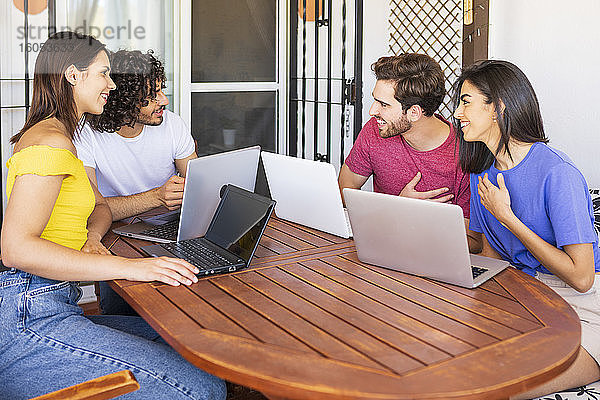 Glückliche junge Männer und Frauen unterhalten sich  während sie mit Laptops am Tisch im Hinterhof sitzen
