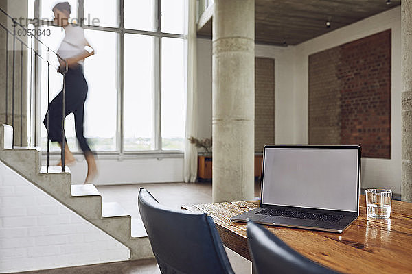 Laptop auf einem Holztisch in einer Loftwohnung mit einer Frau  die im Hintergrund die Treppe hinaufgeht