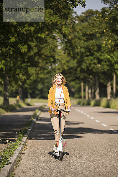 Lächelnde reife Frau fährt elektrischen Roller auf der Straße während des sonnigen Tages