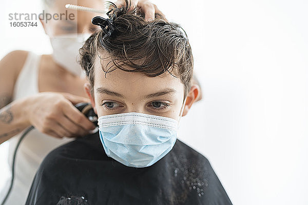 Nahaufnahme eines Friseurs  der einem Jungen mit Maske zu Hause während der Ausgangssperre die Haare schneidet