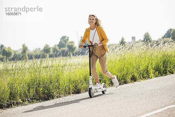 Playful reife Frau Reiten elektrische Push-Roller auf der Straße im Sommer