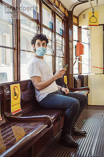 Junger Mann mit Maske  der in der Straßenbahn sitzt und sein Smartphone benutzt