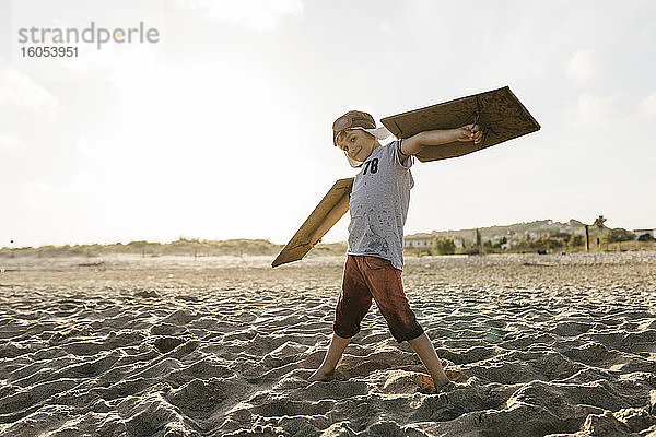 Junge mit Flugzeugflügeln und Mütze  der mit ausgestreckten Armen am Strand steht