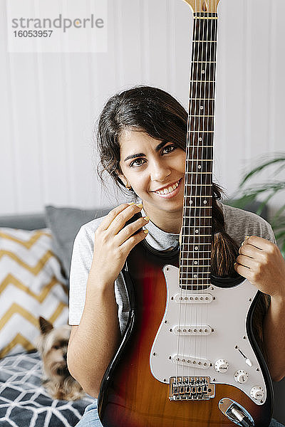 Glückliche Frau  die eine E-Gitarre hält  während sie zu Hause auf dem Sofa sitzt