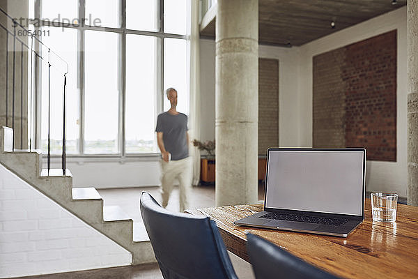 Laptop auf Holztisch in einer Loftwohnung mit Mann im Hintergrund