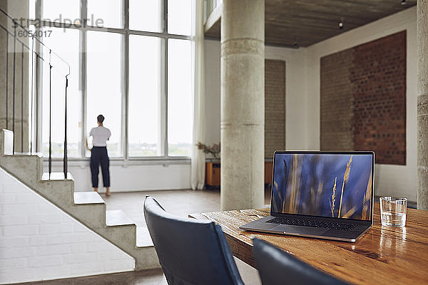 Laptop auf Holztisch in einer Loftwohnung mit Frau am Fenster im Hintergrund
