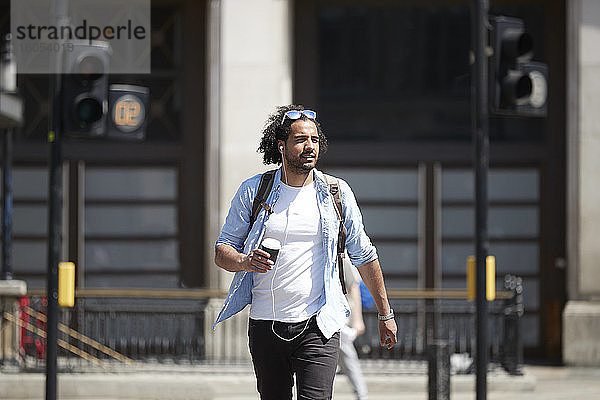 Porträt eines jungen Mannes mit Kaffee zum Mitnehmen  der die Straße überquert  während er mit Kopfhörern Musik hört  London  UK