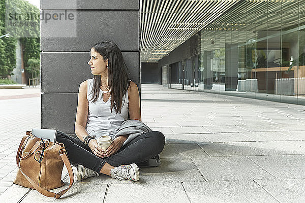 Nachdenkliche Geschäftsfrau  die ein Einwegglas in der Hand hält  während sie auf einer Säule vor einem Bürogebäude sitzt