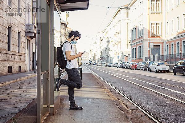 Mann mit Gesichtsmaske  der ein Smartphone benutzt  während er an einem Gebäude in der Stadt steht