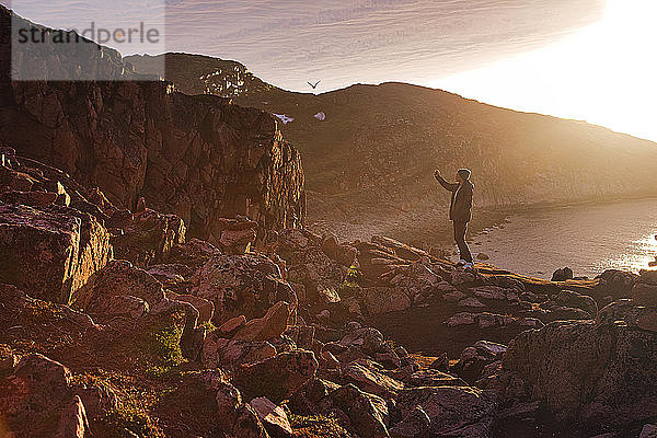 Junger Mann  der ein Selfie macht  während er bei Sonnenuntergang auf einem felsigen Berg in Teriberka  Gebiet Murmansk  Russland  steht