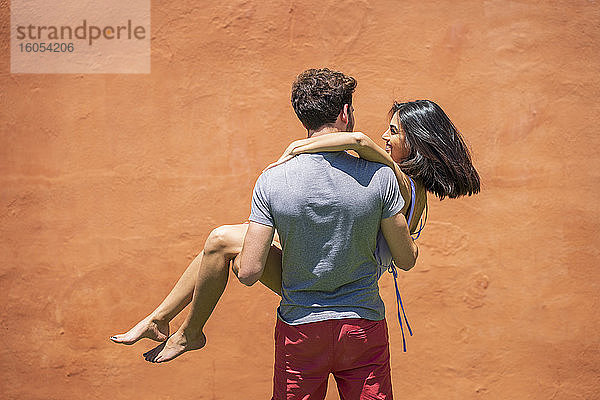 Junger Mann trägt Frau  während er in der Nähe der braunen Mauer im Hinterhof steht