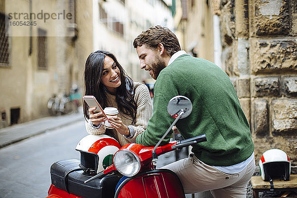 Lächelnde Frau zeigt ihrem Freund das Smartphone  während sie sich auf die Vespa lehnt