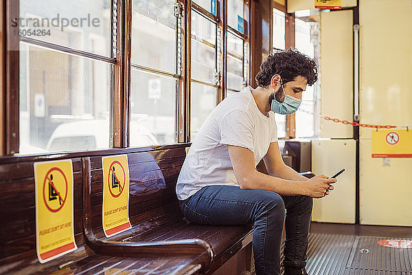 Mann mit Gesichtsmaske benutzt Mobiltelefon in der Straßenbahn
