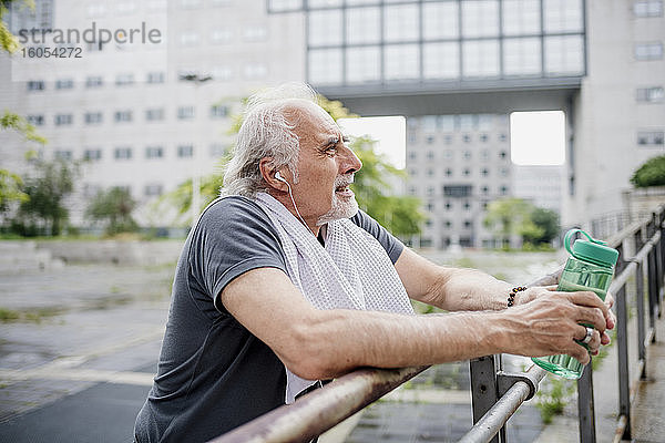 Älterer Mann hält Wasserflasche und hört Musik  während er am Geländer steht