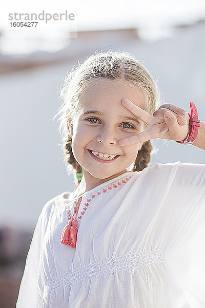Lächelndes süßes Mädchen  das ein Friedenszeichen zeigt  während es im Freien an einem sonnigen Tag steht