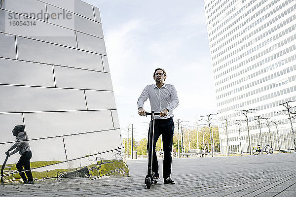 Geschäftsmann mit Kick-Scooter  der sich in der Fassade eines Gebäudes spiegelt