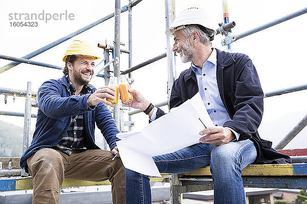 Architekt und Bauarbeiter stoßen mit Getränken an  während sie gegen den klaren Himmel auf der Baustelle sitzen