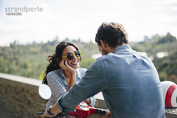 Glückliche Frau mit Sonnenbrille im Gespräch mit ihrem Freund auf einer Vespa sitzend