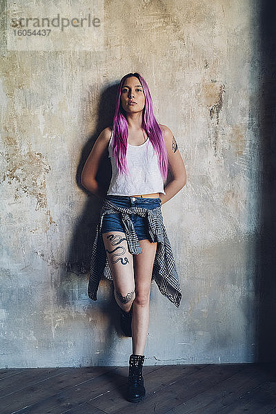 Porträt einer stilvollen jungen Frau mit rosa Haaren  die an einer Wand lehnt