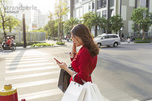 Junge Frau trägt Einkaufstaschen und benutzt ihr Smartphone  während sie auf der Straße steht