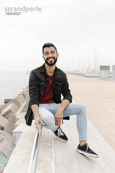 Lächelnder junger Mann sitzt auf einem Geländer gegen den klaren Himmel in der Stadt