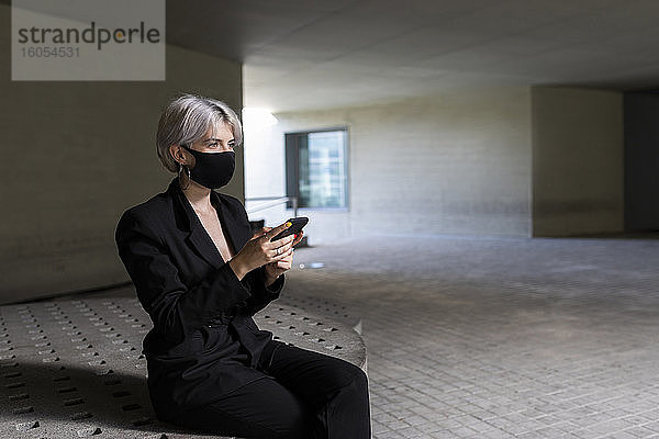 Geschäftsfrau mit Maske  die ein Smartphone benutzt  während sie auf einem Sitz in einem Gebäude sitzt