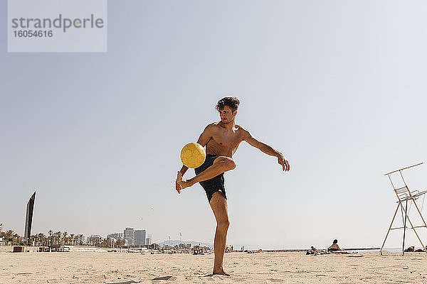 Hemdloser männlicher Sportler spielt mit Ball am Strand gegen klaren Himmel an einem sonnigen Tag