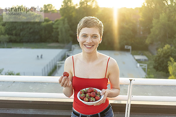 Lächelnde schöne Frau  die Erdbeeren isst  während sie bei Sonnenuntergang auf dem Balkon steht
