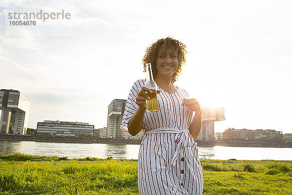 Glückliche Frau mit lockigem Haar  die eine Bierflasche hält  während sie am Fluss in der Stadt steht