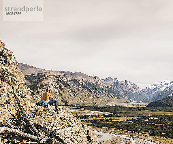 Männlicher Wanderer  der auf einem Felsen sitzend die patagonischen Anden gegen den Himmel betrachtet  Argentinien