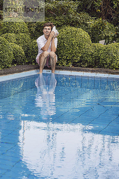 Junger Mann lauscht durch eine Muschel  während er am Pool vor Pflanzen sitzt