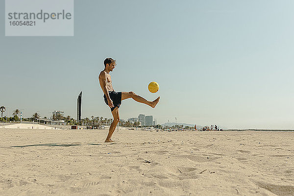 Hemdloser junger Mann spielt mit Ball am Strand gegen klaren Himmel an einem sonnigen Tag