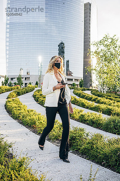 Weiblicher Berufstätiger mit Maske  der ein Smartphone hält  während er auf einem Fußweg in der Stadt spazieren geht
