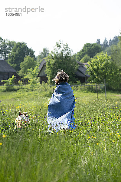 Junge mit Umhang und Hund auf einer Wiese vor klarem Himmel