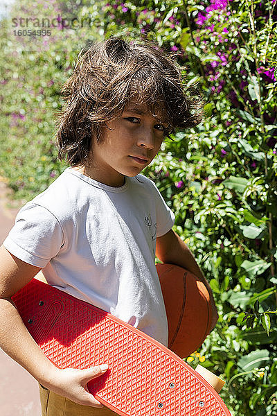Nahaufnahme eines Jungen mit Basketball und Skateboard  der bei Pflanzen im Park steht