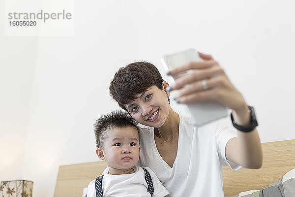 Lächelnde Mutter  die ein Selfie mit ihrem süßen Sohn macht  während sie zu Hause auf dem Bett sitzt