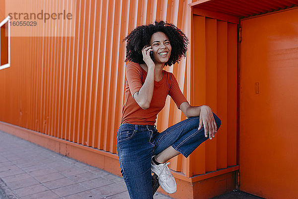 Fröhliche junge Frau  die mit ihrem Smartphone vor einer orangefarbenen Struktur spricht