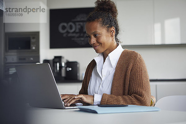 Lächelnde Geschäftsfrau  die am Tisch in der Küche sitzt und einen Laptop benutzt