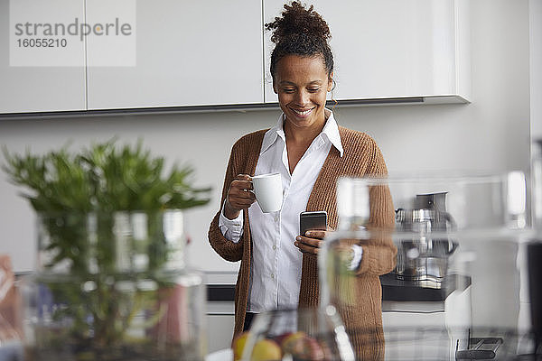 Porträt einer lächelnden Geschäftsfrau  die mit einer Tasse Kaffee in der Küche steht und auf ihr Mobiltelefon schaut