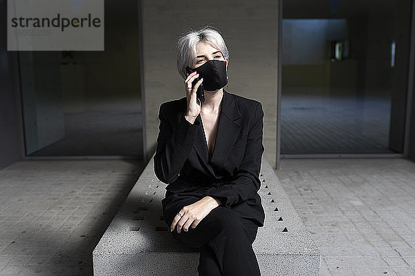 Weibliche Fachkraft mit Maske  die über ein Smartphone spricht  während sie im Büro sitzt