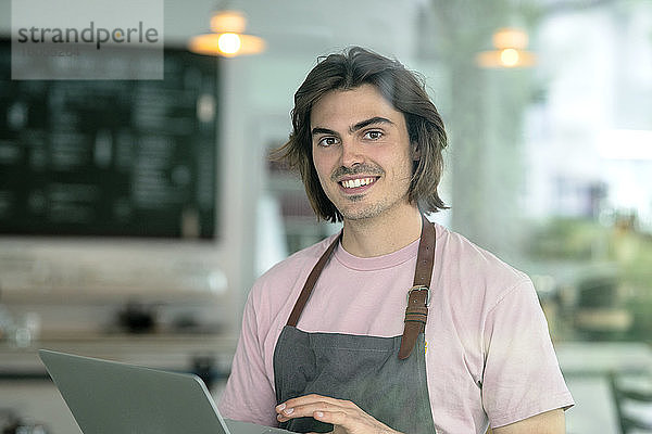 Lächelnder männlicher Besitzer  der einen Laptop benutzt  während er in einem Café steht
