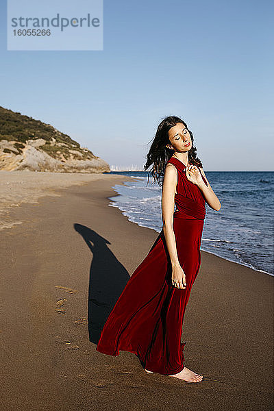 Zarte Frau in rotem Kleid steht am Meer und genießt die Sonne