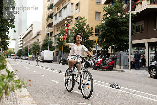 Lächelnder Junge hält Skateboard und fährt Fahrrad auf der Straße