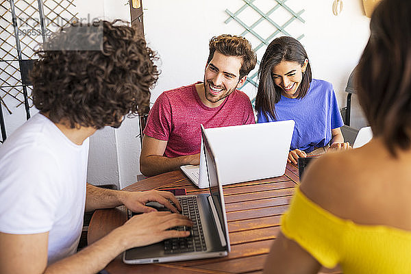 Lächelnde junge Freunde benutzen Laptops  während sie am Tisch im Hinterhof sitzen