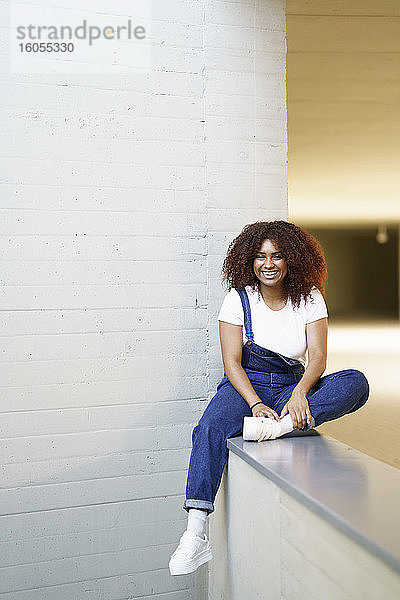 Lächelnde junge Frau mit Afro-Haar sitzt auf einer Stützmauer