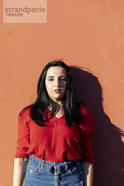 Junge Frau in rotem Hemd steht an einer orangefarbenen Wand in der Stadt an einem sonnigen Tag