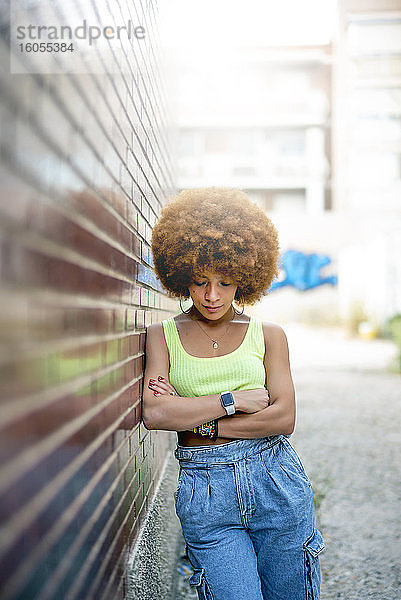 Mittlere erwachsene Frau mit Afro-Haar  die nach unten schaut  während sie an einer Backsteinmauer in der Stadt steht