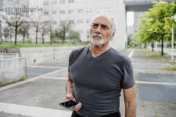 Älterer Mann hält Smartphone und schaut weg  während er auf einem Fußweg in der Stadt steht