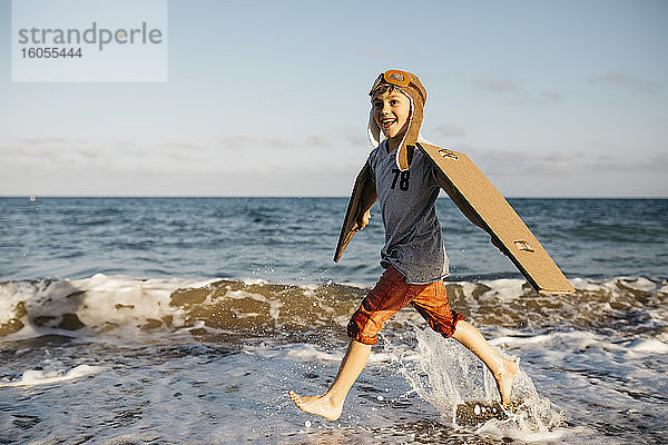 Junge mit Flugzeugflügeln und Mütze spritzt beim Laufen am Strand