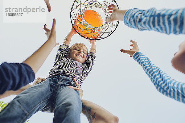 Glücklicher Junge wirft Basketball in den Korb  während er mit seiner Familie gegen den klaren Himmel spielt